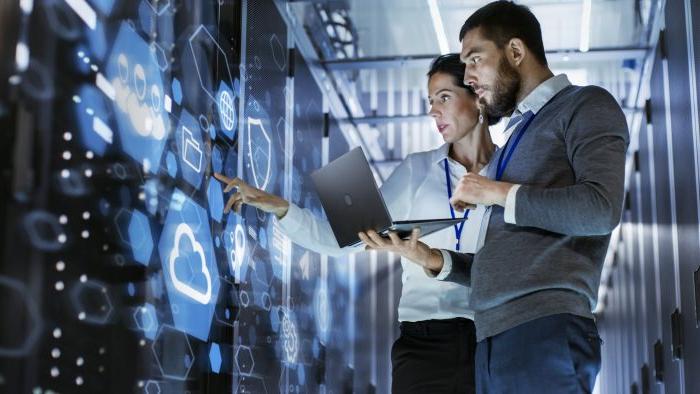 A female technology manager stands utilizing a touch screen console while her male counterpart holds a laptop at the ready.